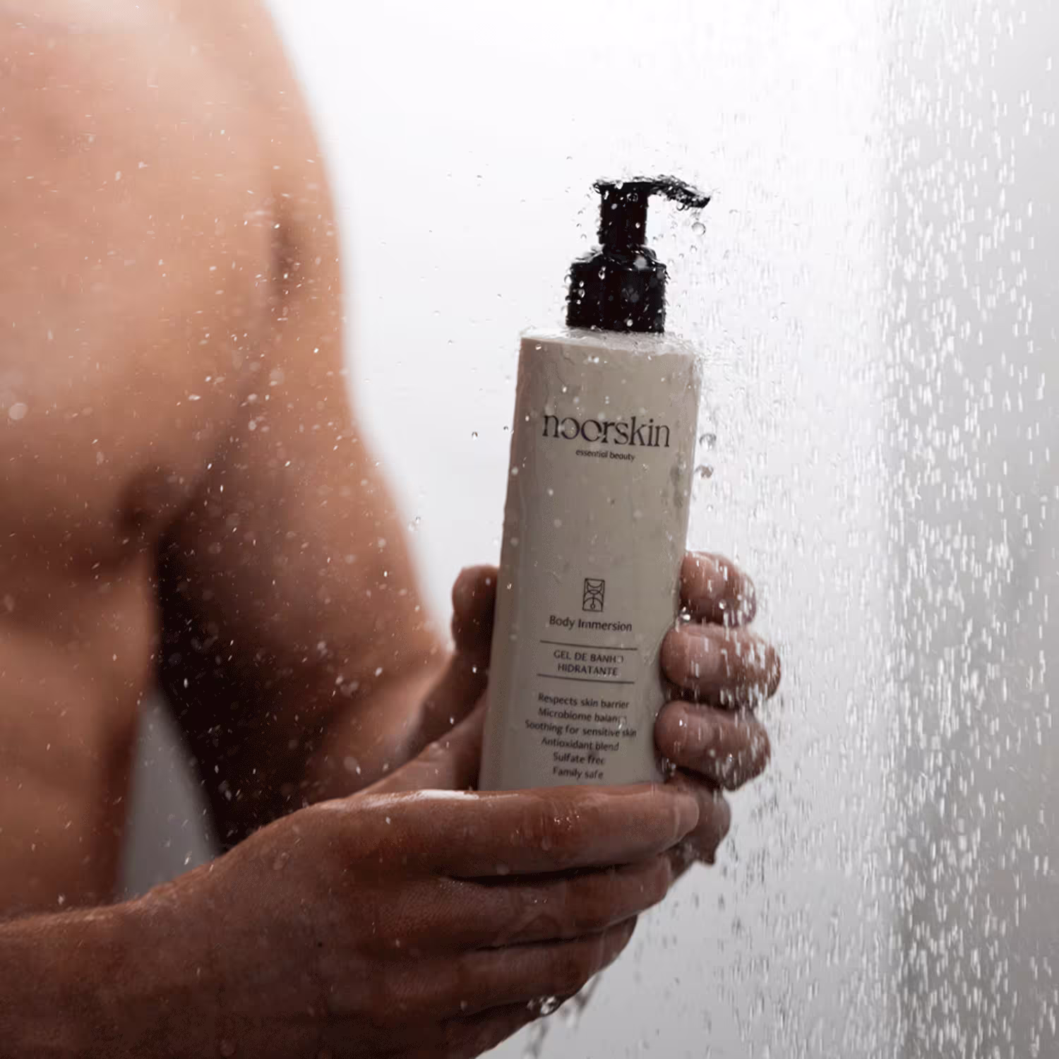 Mão masculina segura o Gel de Banho Noorskin Essential Beauty Body Immersion, sendo molhado pela água do chuveiro