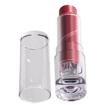 A imagem mostra a tampa da embalagem de balm, semelhante a uma tampa de batom, transparente e cilíndrica. Ao lado, a embalagem do balm, com o produto de cor rosada com a palavra KIND escrito na embalagem.