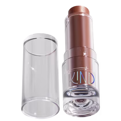 A imagem mostra a tampa da embalagem de balm, semelhante a uma tampa de batom, transparente e cilíndrica. Ao lado, a embalagem do balm, com o produto de cor marrom com a palavra KIND escrito na embalagem.