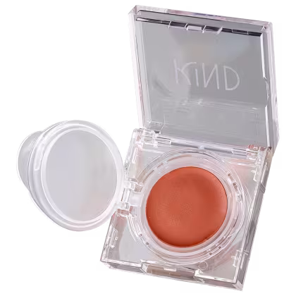 A imagem mostra a embalagem de um blush rosa claro. Em formato redondo, dentro de uma caixinha de acrílico transparente. A caixa está aberta e na parte superior, tem o nome da marca em destaque.