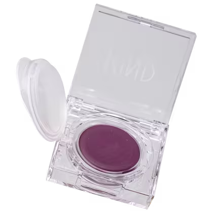 A imagem mostra a embalagem de um blush roxo. Em formato redondo, dentro de uma caixinha de acrílico transparente. A caixa está aberta e na parte superior, tem o nome da marca em destaque.