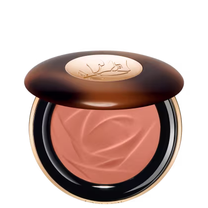 A imagem traz um sérum bronzeador compacto em uma embalagem. A tampa superior é marrom com alguns detalhes mais claros e tons mais escuros. O logotipo da marca aparece gravado em dourado no centro da tampa e o produto possui uma cor bege.