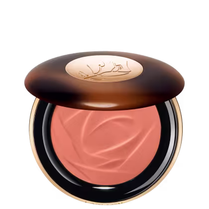 A imagem traz um sérum bronzeador compacto em uma embalagem. A tampa superior é marrom com alguns detalhes mais claros e tons mais escuros. O logotipo da marca aparece gravado em dourado no centro da tampa e o produto possui uma cor rosada.