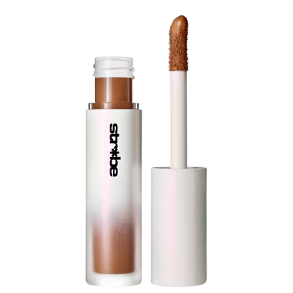 O produto de beleza apresentado é um corretivo líquido da marca Strobe, em embalagem branca e marrom, com aplicador em formato de pincel. O frasco possui um rótulo com o nome da marca em destaque.