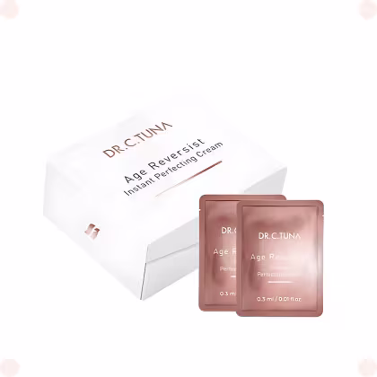 Dentro da caixa, duas embalagens marrons achatadas contendo 0,3 ml cada do creme anti-idade. As embalagens apresentam o nome da marca e do produto impresso em letras douradas.