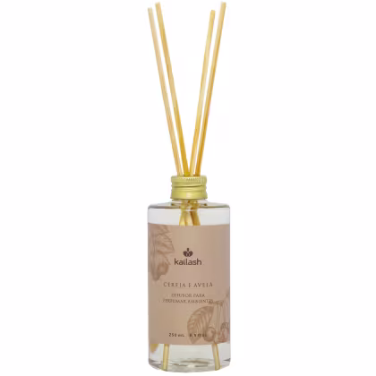 Difusor de ambiente Kailash com aroma de cereja e avelã, em frasco de vidro transparente com rótulo bege e tampa dourada, contendo varetas de madeira para dispersão do aroma.