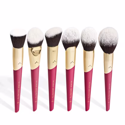 A imagem mostra seis pincéis de maquiagem, lado a lado, com cabo vermelho e dourado, e cerdas pretas com ponta branca. Na parte vermelha, logo da marca e linha do produto escrito em dourado.