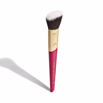 A imagem mostra um pincel de maquiagem, visto de um ângulo um pouco mais elevado, sem ser exatamente de frente. Com cabo vermelho e dourado, e cerdas pretas com ponta branca. Na parte vermelha, logo da marca e linha do produto escrito em dourado.