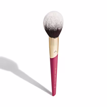 A imagem mostra um pincel de maquiagem, visto de um ângulo um pouco mais elevado, sem ser exatamente de frente. Com cabo vermelho e dourado, e cerdas pretas com ponta branca. Na parte vermelha, logo da marca e linha do produto escrito em dourado.