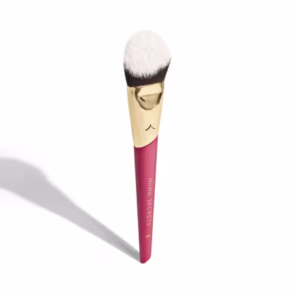 A imagem mostra um pincel de maquiagem, visto de um ângulo um pouco mais elevado, sem ser exatamente de frente. Com cabo vermelho e dourado, e cerdas pretas com ponta branca. Na parte vermelha, logo da marca e linha do produto escrito em dourado.