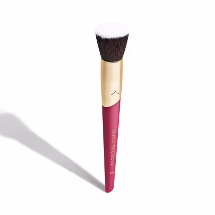 A imagem mostra um pincel de maquiagem, visto de um ângulo um pouco mais elevado, sem ser exatamente de frente. Com cabo vermelho e dourado, e cerdas pretas com ponta branca. Na parte vermelha, logo da marca e linha do produto escrito em dourado.