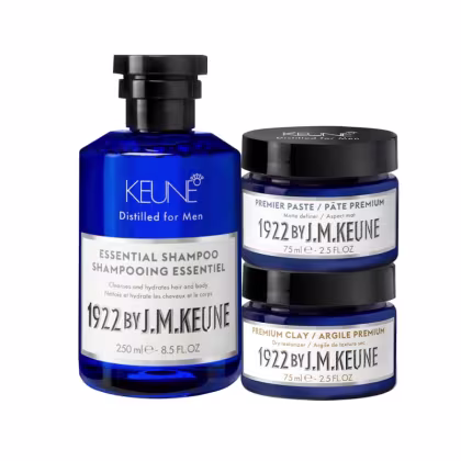 Produtos de cuidados capilares da marca Keune, incluindo um frasco de shampoo essencial de 250 ml e dois potes de 75 ml, um de pasta premium e outro de argila premium, todos em embalagens de vidro azul com rótulos brancos e pretos.