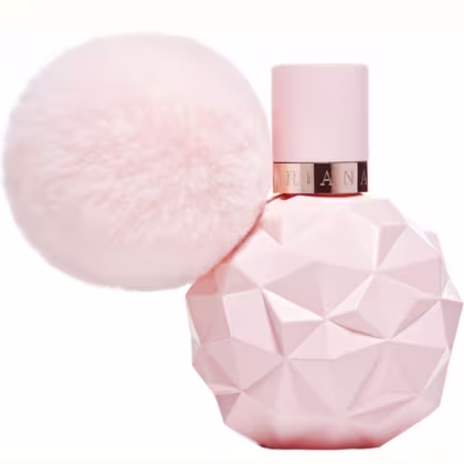 Frasco de perfume Ariana Grande em formato geométrico rosa, com tampa rosada e detalhes em metal dourado. Acompanha um elemento decorativo esférico e felpudo em tons de rosa.