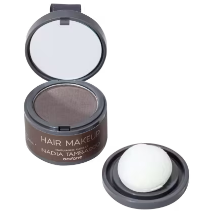 A imagem mostra a maquiagem para cabelos, é um pó compacto em uma embalagem redonda preta com espelho na tampa espelhada. O pó tem uma cor marrom escuro. Também há um pequeno compartimento separado com um aplicador de esponja branca.