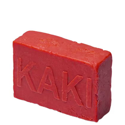 Barra de sabonete retangular de cor laranja viva. A palavra "KAKI" está gravada em relevo na superfície com letras maiúsculas e em negrito. A textura apresenta pequenas rachaduras e irregularidades, sugerindo um aspecto rústico e artesanal.