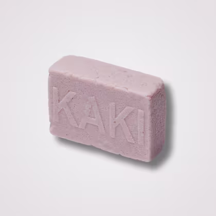 Barra de sabonete rosa claro em formato retangular. Tem textura levemente áspera e granulada. O nome "KAKI" está gravado em letras maiúsculas na superfície do sabonete. O tom rosado suave e a aparência rústica sugerem que pode ser um sabonete artesanal.