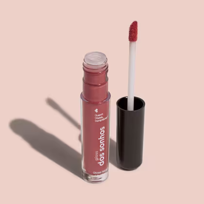Frasco cilíndrico transparente contendo um gloss labial marrom. Rótulo na embalagem com o nome do produto "Gloss dos Sonhos" e a descrição "Quem disse, berence?". Ao lado, a tampa preta do produto, que acompanha um aplicador de espuma.