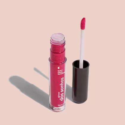 Frasco cilíndrico transparente contendo um gloss labial rosa brilhante Rótulo na embalagem com o nome do produto "Gloss dos Sonhos" e a descrição "Quem disse, berence?". Ao lado, a tampa preta do produto, que acompanha um aplicador de espuma rosa.