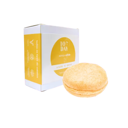 O produto apresentado é o shampoo sólido Ucuuba & Alecrim da marca Today Natural Cosmetics. Mostra uma caixa branca com detalhes em amarelo. Ao lado da caixa, há o shampoo sólido em formato de barra arredondada de cor amarelada.