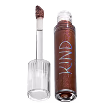 A imagem mostra a tampa da embalagem do gloss, com o aplicador virado pra cima, semelhante a uma tampa de batom, transparente e cilíndrica. Ao lado, o frasco do gloss, com o produto de cor marrom escuro e com a palavra KIND escrito na embalagem.