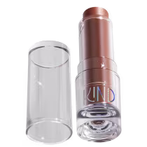 A imagem mostra a tampa da embalagem de balm, semelhante a uma tampa de batom, transparente e cilíndrica. Ao lado, a embalagem do balm, com o produto de cor marrom com a palavra KIND escrito na embalagem.