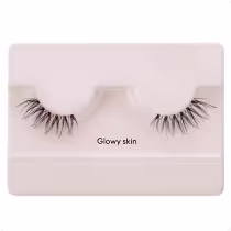 A imagem mostra os cílios postiços em um estojo plástico claro. O estojo tem uma cor nude claro e exibe um par de cílios postiços pretos.