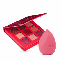 A imagem mostra um kit com paleta de sombras e uma esponja. A paleta é toda vermelha com 9 cores sendo tons de marrom, roxo e dourado. Já a esponja tem formato triangular e cor rosa.