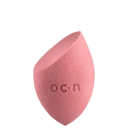 Esponja de maquiagem de cor rosa, com formato assimétrico e textura macia, com o logotipo "O.C.N." impresso na superfície.