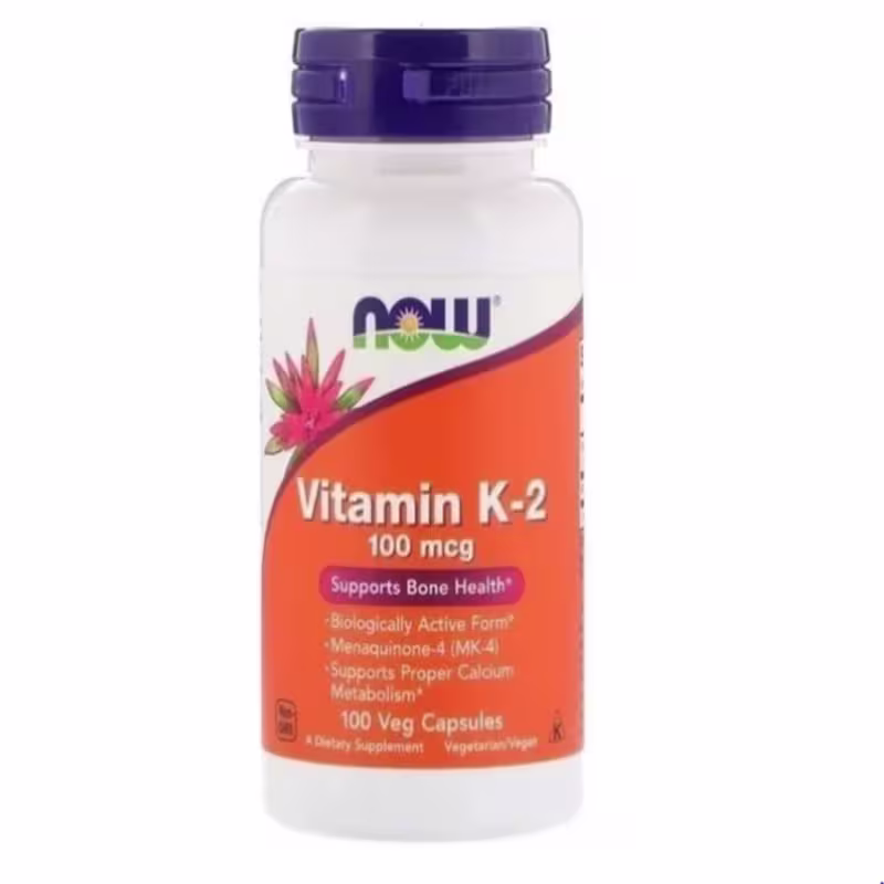 Este é um suplemento vitamínico da marca Now Foods, em um pote branco com tampa azul. O rótulo laranja destaca que é Vitamina K-2 na dosagem de 100 mcg. Menciona que suporta a saúde óssea e o metabolismo adequado do cálcio. O frasco contém 100 cápsulas