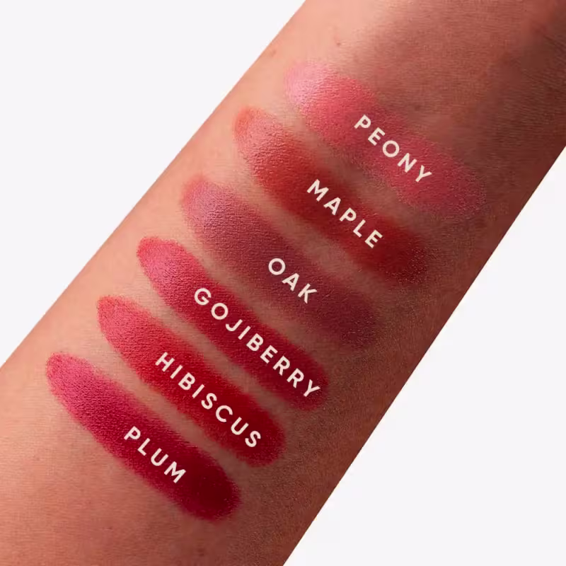 A imagem mostra uma série de amostras da cor do blush em diferentes tons de vermelho, variando de um rosa claro a um vermelho escuro. Os nomes dos tons estão impressos sobre cada amostra, incluindo "Peony", "Maple Oak", "Goji Berry" e "Hibiscus Plum".