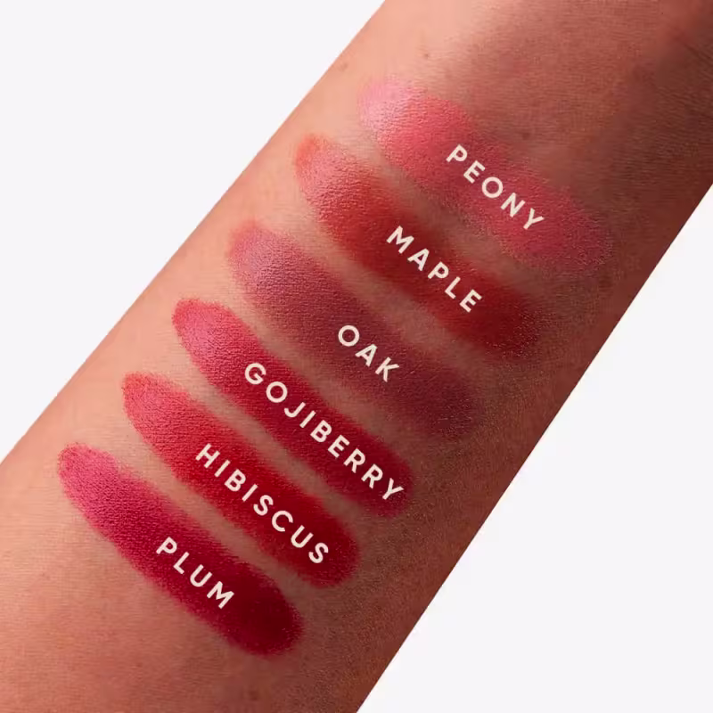 A imagem mostra uma série de amostras da cor do blush em diferentes tons de vermelho, variando de um rosa claro a um vermelho escuro. Os nomes dos tons estão impressos sobre cada amostra, incluindo "Peony", "Maple Oak", "Goji Berry" e "Hibiscus Plum".