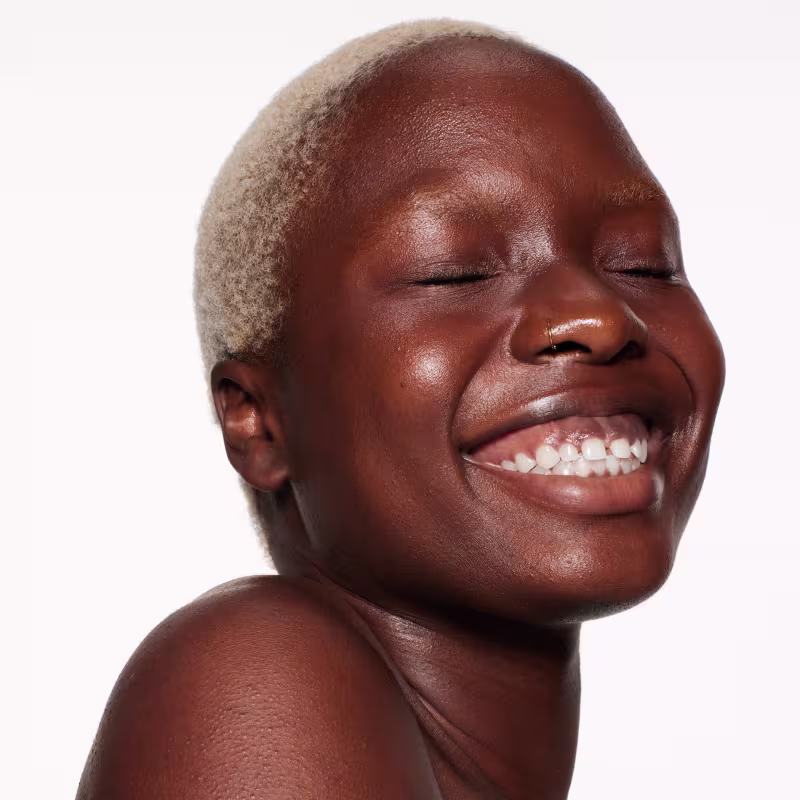 Esta imagem mostra um close-up do rosto sorridente de uma pessoa negra. A pessoa tem pele escura brilhante, olhos fechados e um largo sorriso revelando dentes brancos. A expressão é alegre e descontraída, transmitindo uma sensação de felicidade.