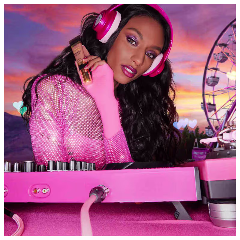 A imagem mostra uma mulher super produzida, com fone de ouvido e uma mesa de dj, ambos de cor rosa, segurando uma embalagem da base próxima ao rosto.