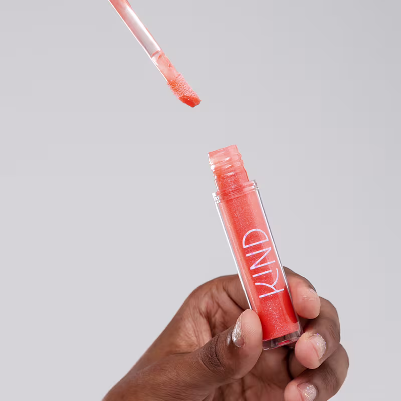 A imagem mostra a embalagem do gloss, aberta, sendo segurado por uma mão negra com unhas curtas.