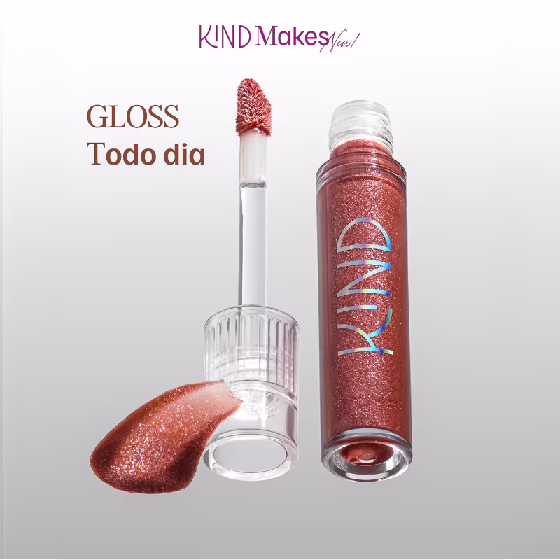 A imagem mostra a tampa da embalagem do gloss com o aplicador virado pra cima, semelhante a uma tampa de batom, transparente, cilíndrica, com uma amostra de cor. Ao lado, o frasco do gloss, com o produto de cor marrom e palavra KIND escrito na embalagem.