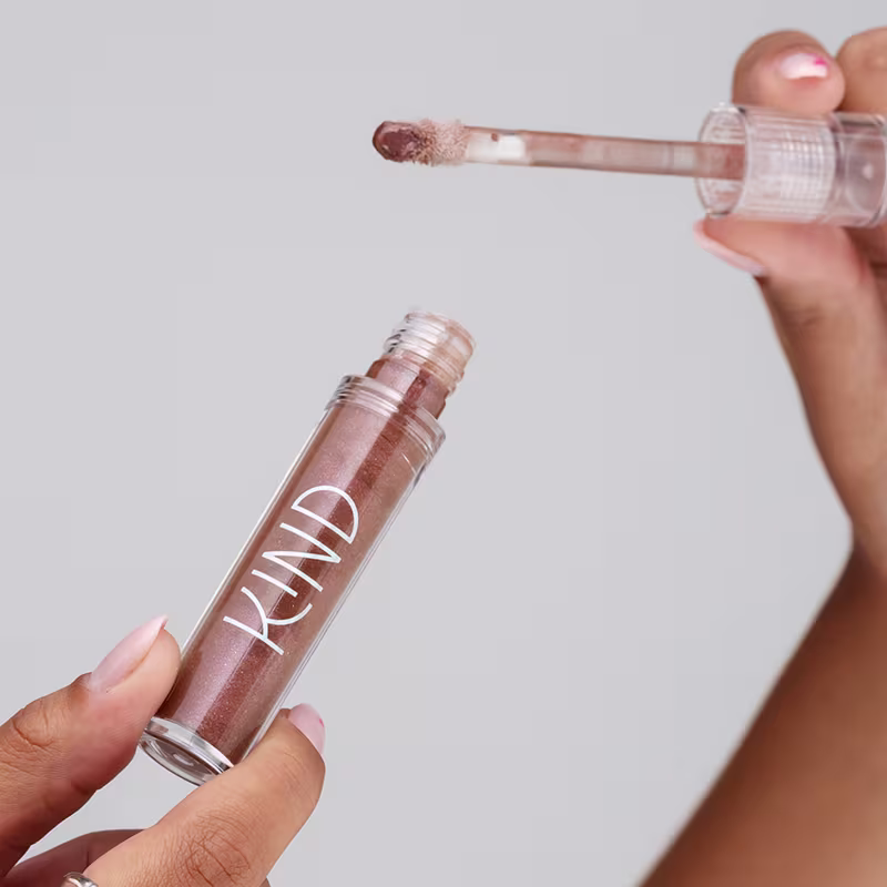 A imagem mostra a embalagem do gloss, aberta, sendo segurado por uma mão com unhas claras e anéis prateados.