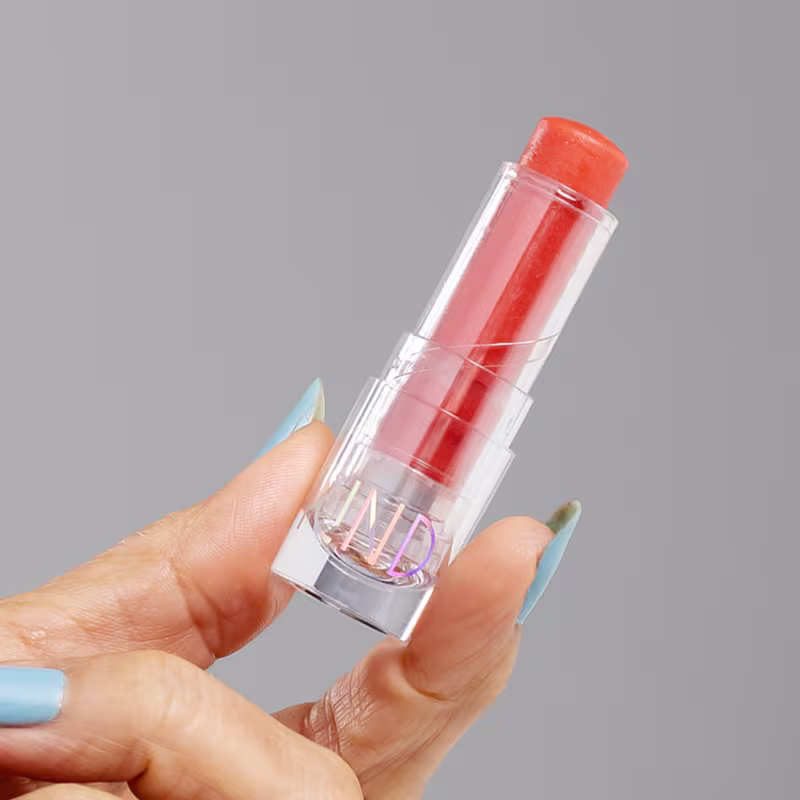 A imagem mostra a embalagem do balm, aberta, sendo segurado por uma mão branca com esmalte azul.