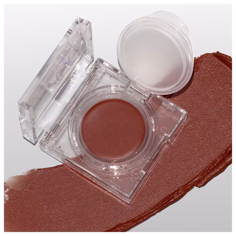 A imagem mostra a embalagem do blush cremoso aberta, numa superfície branca com um pouco do blush esparramado pela superfície, mostrando a cor e a textura cremosa.