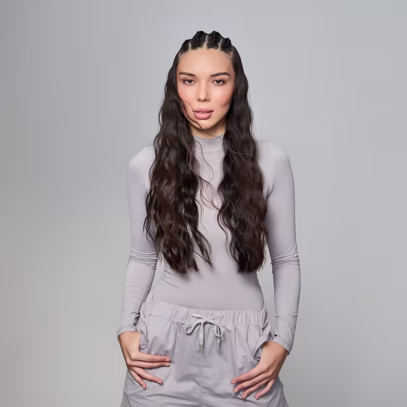 A imagem mostra uma mulher com cabelos longos castanhos escuros e ondulados. Ela está usando um conjunto de blusa de manga longa e calça na cor cinza em tom claro.