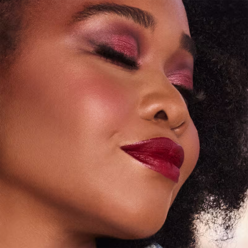 A imagem mostra uma mulher negra de cabelos cacheados com uma bela maquiagem em tons de vermelho e rosa. A sombra de olhos é metalizada com tons de vermelho e rosa. O batom é vermelho intenso e brilhante. A pele tem um lindo bronzeado natural e saudável.