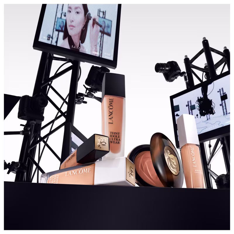 A imagem mostra alguns produtos de maquiagem Lancôme. Frascos de base líquida, na cor bege, embalagens cilíndricas com tampas pretas e brancas, com rótulos pretos e o logotipo da marca. Ao fundo alguns monitores como em um estúdio de gravação.