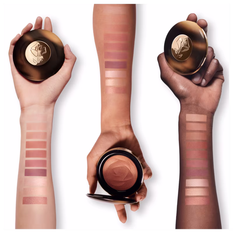 A imagem trás três embalagens com o bronzer. Elas têm formatos cilíndricos e são segurados por mãos femininas de diferentes tons de pele. No antebraço estão as tonalidades do bronzer e como eles ficam em cada pele.