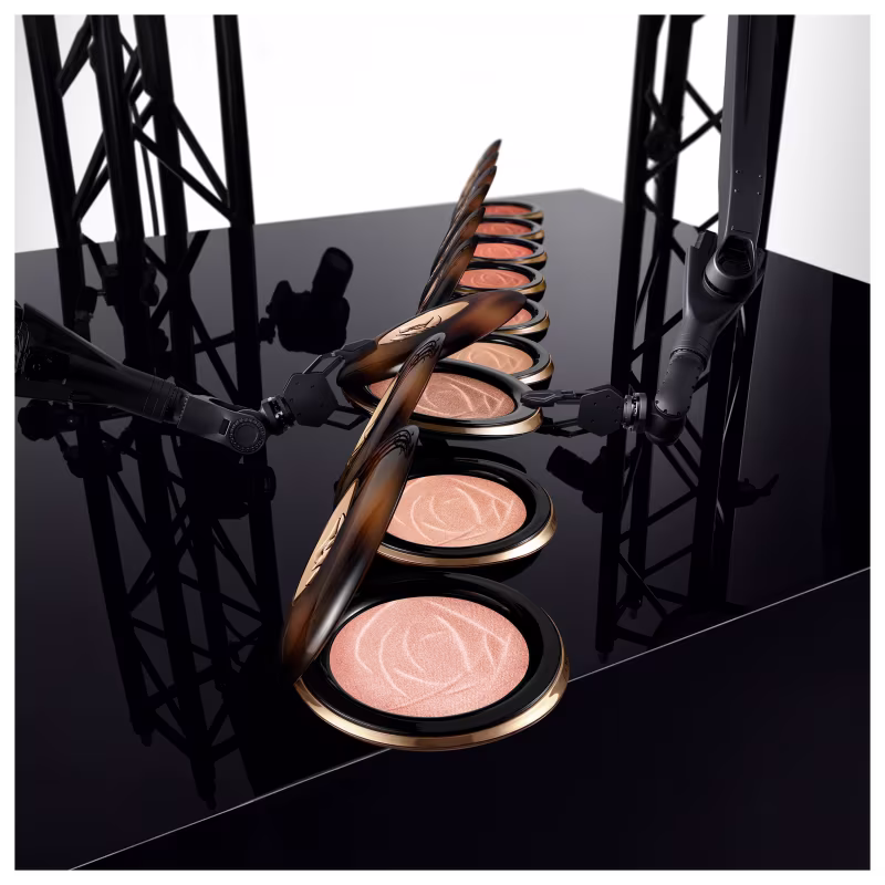 A imagem mostra bronzers de diferentes tonalidades dispostos em uma fileira sobre uma superfície escura, com estruturas metálicas pretas ao fundo. As cores predominantes são preto, dourado e tons de rosa claro.