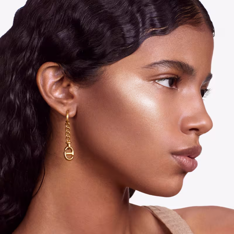 Esta é uma imagem de uma mulher com pele bronzeada e traços delicados, promovendo um produto de beleza. Ela tem cabelos escuros e ondulados, olhos bem delineados com maquiagem e lábios levemente rosados. Usa um brinco dourado com um design de elos.