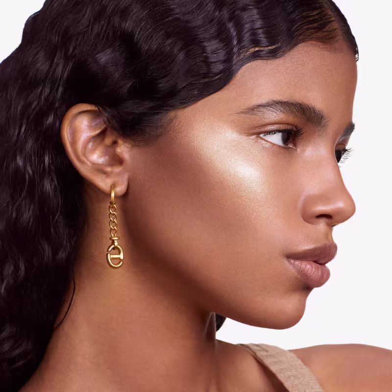 Esta é uma imagem de uma mulher com pele bronzeada e traços delicados, promovendo um produto de beleza. Ela tem cabelos escuros e ondulados, olhos bem delineados com maquiagem e lábios levemente rosados. Usa um brinco dourado com um design de elos.