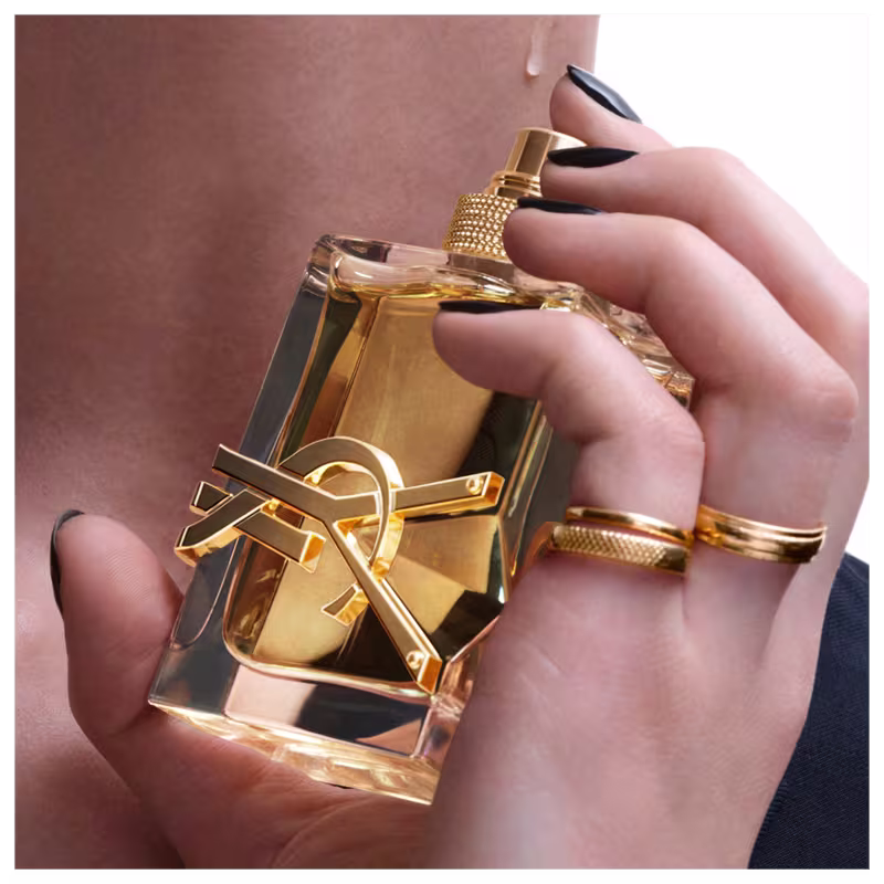 A imagem mostra uma mão feminina com unhas pretas e anéis dourados aplicando o perfume próxima ao pescoço.