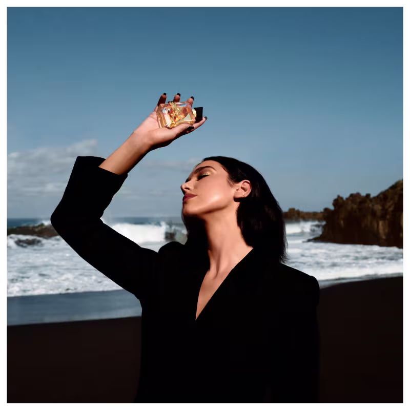 A imagem mostra a cantora Dua Lipa, uma mulher jovem branca de cabelos pretos curtos, segurando o perfume. Ela está com um terno preto e ao fundo é possível ver uma praia.