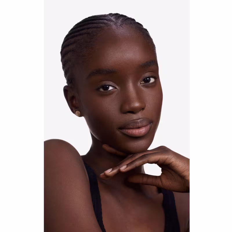 Esta imagem apresenta uma jovem mulher negra com um penteado de tranças finas presas rente à cabeça. Ela tem olhos castanhos, lábios delineados e pele em um tom profundo de marrom. Sua expressão é serena, com uma mão delicadamente apoiada no pescoço.