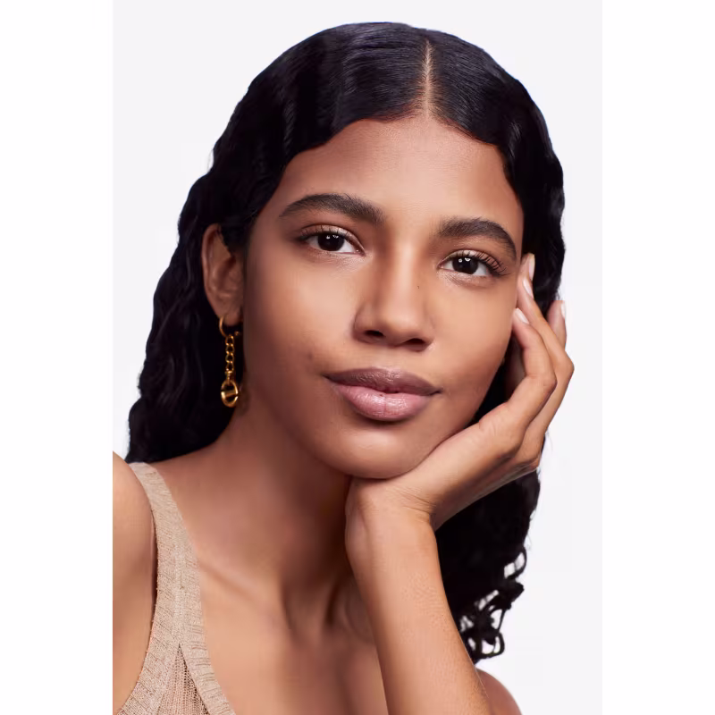 Esta imagem apresenta uma modelo jovem com traços étnicos. Ela tem pele bronzeada, olhos castanhos marcantes e lábios cheios e rosados. Seus cabelos são negros e ondulados. Ela usa brincos dourados e toca suavemente o rosto com uma das mãos.