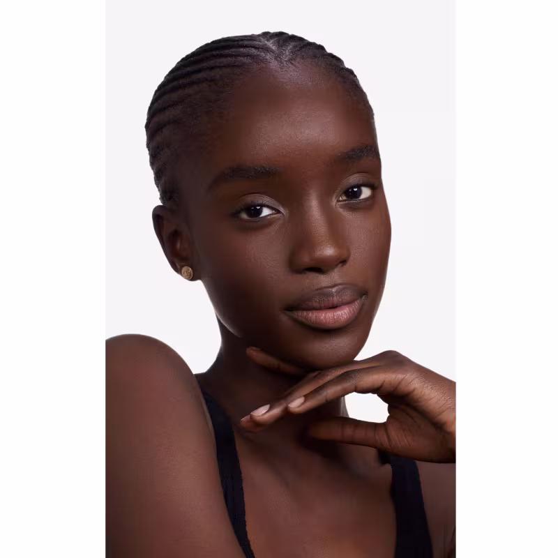Esta imagem apresenta uma jovem mulher negra com um penteado de tranças finas presas rente à cabeça. Ela tem olhos castanhos, lábios delineados e pele em um tom profundo de marrom. Sua expressão é serena, com uma mão delicadamente apoiada no pescoço.